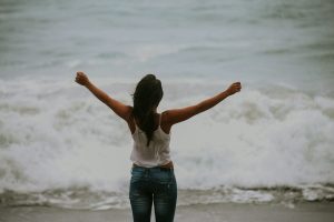 woman in white spaghetti strap top standing on the seashore extending her arms