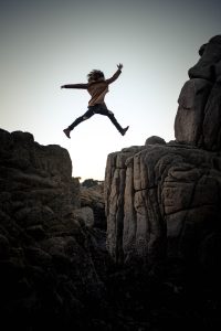 child jumping to a big rock under gray and white sky during daytime courageously